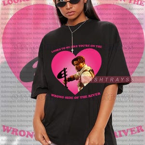 May include: Black unisex t-shirt featuring a pink heart graphic with an image of a man holding a prop. The text "LOOKS TO ME LIKE YOU'RE ON THE WRONG SIDE OF THE RIVER" is printed on the shirt.