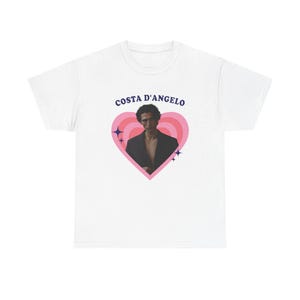 Può includere: T-shirt bianco con una grafica di una persona all'interno di un cuore rosa, con la scritta "COSTA D'ANGELO" sopra. La grafica include accenti di stelle blu. La persona nella grafica indossa un blazer nero.