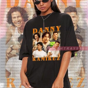 May include: Black t-shirt with a graphic print of a group of men with the text "DANNY RAMIREZ" in orange. The model is wearing a size 2XL shirt.