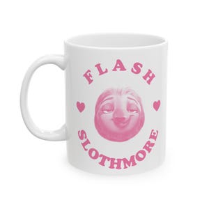 Puede incluir: Taza de cerámica blanca con una ilustración rosa de la cara de un perezoso, rodeada por las palabras "FLASH SLOTHMORE" en rosa, con dos corazones rosas. La taza tiene un asa curva.
