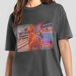 Op de afbeelding: Een antracietgrijs t-shirt met korte mouwen en een rechthoekige grafische print van een oranje paardachtig personage in een kamer. Het personage heeft een paarse hartvormige vlek op het achterwerk. De kamer bevat een boekenplank, een roze tafel en een raam.
