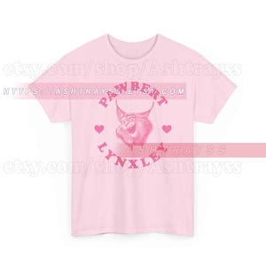 May include: A pale pink t-shirt with a cartoon lynx design. The design includes the text "PAWBERT LYNXLE" in pink, a lynx illustration, and heart shapes. The Etsy shop URL is visible.