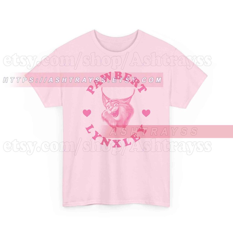 May include: A pale pink t-shirt with a cartoon lynx design. The design includes the text "PAWBERT LYNXLE" in pink, a lynx illustration, and heart shapes. The Etsy shop URL is visible.