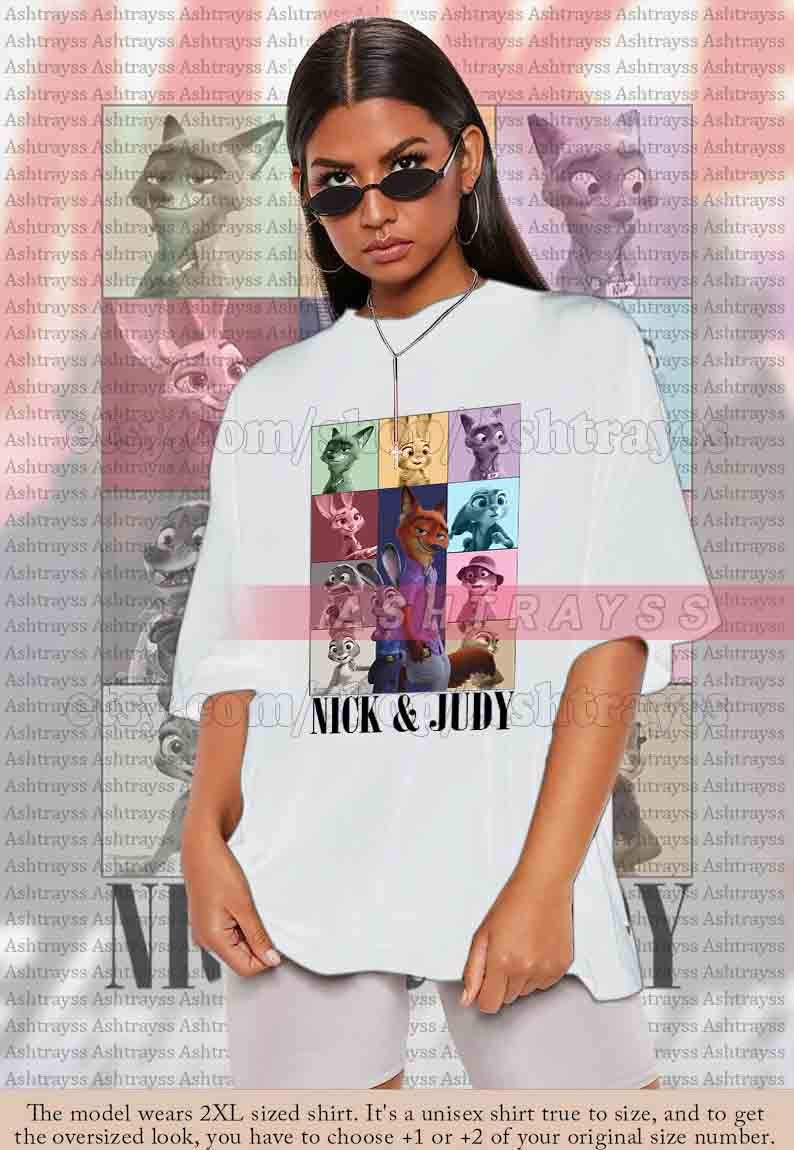 May include: White unisex t-shirt featuring a colorful graphic of characters from the movie "Zootopia", with the text "NICK & JUDY". The shirt is oversized and has a crew neck.