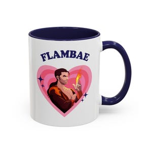 Puede incluir: Taza de cerámica blanca con interior y asa azul marino. La taza presenta un gráfico de un hombre con una llama en el dedo dentro de un corazón rosa, con la palabra "FLAMBAE" encima. La taza es un artículo de novedad.