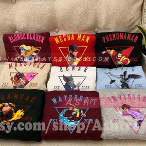 May include: A collection of nine sweatshirts in various colors, each featuring a superhero graphic and text. The sweatshirts display names like "Blonde Blazer" and "Mecha Man," with "EST. 2025" printed below the graphics. The sweatshirts are displayed on a beige couch.