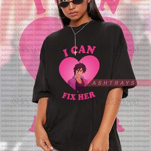 Könnte beinhalten: Schwarzes Unisex-T-Shirt mit dem pinkfarbenen Text "I CAN FIX HER" und einer Grafik einer Figur in einem rosa Herzen. Das übergroße Shirt zeigt auch den Markennamen "ASHTRAYS".