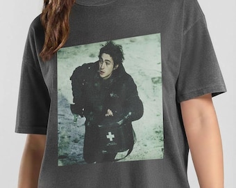 May include: A charcoal gray short-sleeve t-shirt featuring a rectangular graphic print of a person in a dark jacket carrying a black medical bag with a white cross symbol. The person has dark, messy hair and is looking toward the upper left.