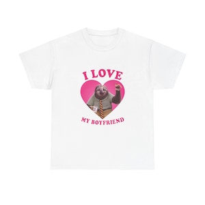 Puede incluir: Camiseta blanca con un perezoso de dibujos animados dentro de un corazón rosa. El perezoso sostiene una taza de café y señala hacia arriba. El texto "I LOVE MY BOYFRIEND" está impreso encima y debajo del corazón en rosa.