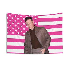 May include: A rectangular wall tapestry featuring a stylized American flag pattern in shades of hot pink and white, with a portrait of a person with short dark hair wearing a brown collared shirt and a matching brown coat layered over it.