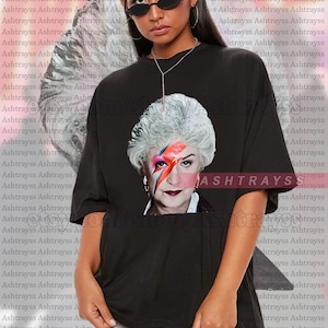 May include: Black unisex t-shirt featuring a graphic of a woman with white hair and a lightning bolt across her face. The shirt has the word "ASHTRAYSS" printed on it. The model is wearing a 2XL shirt.