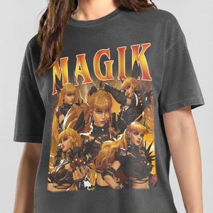 Könnte beinhalten: Ein anthrazitfarbenes T-Shirt mit einem Grafikdruck, der das Wort MAGIK in fetten orangefarbenen und gelben Buchstaben zeigt. Das Design zeigt mehrere Illustrationen einer blonden Figur in dunkler Rüstung vor einem gelb-orangen Blitzhintergrund.