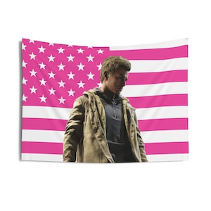 Peut inclure: Une tapisserie murale rectangulaire représentant un drapeau américain rose et blanc. Une personne se tient devant le drapeau, vêtue d'une veste marron et d'un sweat à capuche gris. La tapisserie est probablement en tissu.