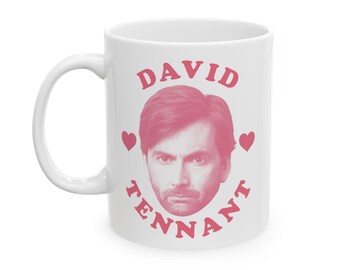 May include: White ceramic mug featuring a pink image of David Tennant, with the name "DAVID TENNANT" in pink lettering. Two pink hearts flank the image. The mug has a white handle.