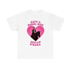 以下が含まれることがあります： 中央にピンクのハートのグラフィックが描かれた白いTシャツ。ハートの中には黒い馬に乗った人物のイラストがあります。ハートの上にはピンク色で「SAVE A HORSE, RIDE」、下には「DECLAN O'HARA」という文字がプリントされています。