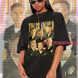 May include: A black t-shirt with a graphic print of a group of men in police uniforms. The text "TIM BRADFORD" is printed in gold letters above the image. The t-shirt is oversized and the model is wearing a size 2XL.