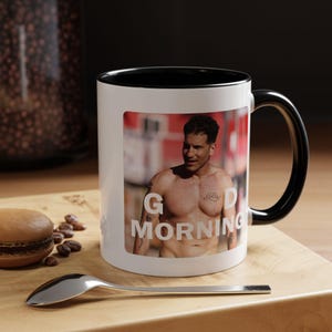 Op de afbeelding: Een witte keramische mok met een zwarte handgreep en binnenrand. De mok is voorzien van een fotoprint van een man met ontbloot bovenlijf en tatoeages, met de witte tekst "GOOD MORNING" over de afbeelding heen. De mok is cilindervormig.