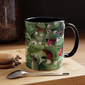 May include: A ceramic mug with a black interior and handle, featuring a repeating pattern of cartoon green frogs with large eyes and expressive faces. The frogs are depicted in various poses, some with tongues sticking out, covering the exterior surface.