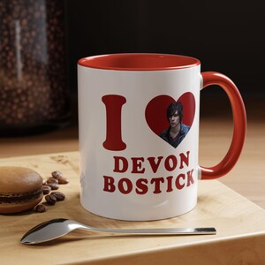May include: A white ceramic mug with a red interior and handle. The front features the red text I, a red heart containing a photographic portrait of Devon Bostick, and the red text DEVON BOSTICK printed in a bold, sans-serif font below the heart.