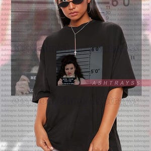 May include: Black unisex t-shirt with a framed image of a woman holding a sign that reads "METRO POLICE FRAN FINE". The shirt also has a height chart graphic and the text "ASHTRAYSS".