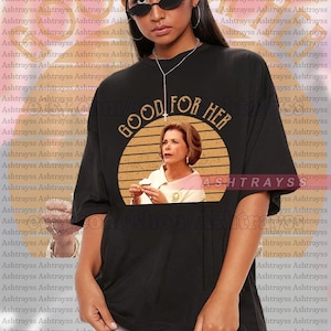 Puede incluir: Camiseta unisex negra con el texto "GOOD FOR HER" en letras doradas. La camiseta presenta un gráfico circular con la imagen de una mujer. La marca "ASHTRAYS" está impresa en la camiseta.