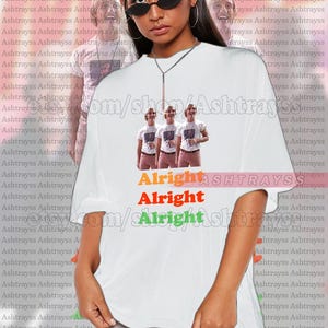 May include: White unisex t-shirt featuring a graphic of three people and the text "Alright Alright Alright" in orange, red, and green. The shirt is oversized and has a crew neck.