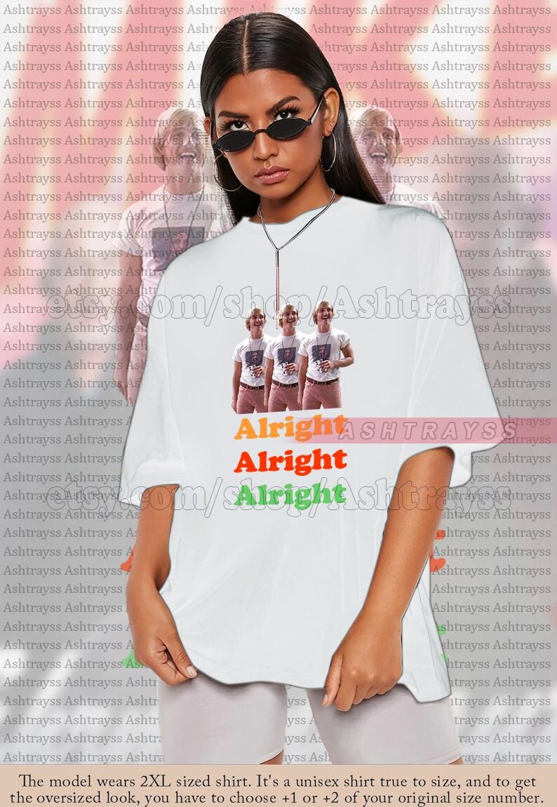 May include: White unisex t-shirt featuring a graphic of three people and the text "Alright Alright Alright" in orange, red, and green. The shirt is oversized and has a crew neck.
