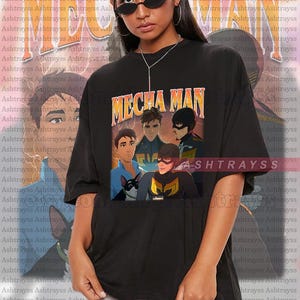 以下が含まれることがあります： 黒色のユニセックスTシャツ。アニメのキャラクターのグラフィックとオレンジと黄色の「MECHA MAN」の文字がプリントされています。 ゆったりとしたサイズで、クルーネックです。