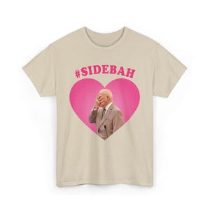 May include: A beige short-sleeved t-shirt featuring a large pink heart graphic on the chest. Inside the heart is a photograph of a person in a suit covering their face with one hand. Above the heart, the text #SIDEBAH is printed in pink.