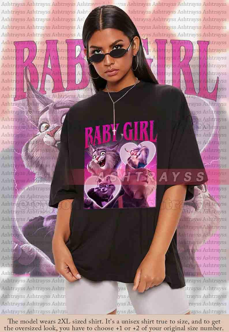May include: Black unisex t-shirt featuring a graphic with the text "BABY GIRL" in pink, along with cartoon cat illustrations. The shirt is oversized and has short sleeves.