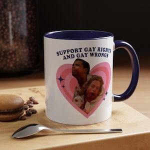 May include: White ceramic mug with a navy blue handle and rim. The mug features the text "SUPPORT GAY RIGHTS AND GAY WRONGS" above a pink heart with two people inside. The mug is on a wooden surface.