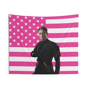 Puede incluir: Un tapiz rectangular con un diseño de bandera estadounidense rosa y blanca. Una persona con un atuendo negro se encuentra frente a la bandera. El tapiz parece ser de tela.