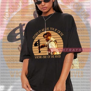 Puede incluir: Camiseta negra unisex con un diseño gráfico circular de un hombre sosteniendo un arma, con el texto "LOOKS TO ME LIKE YOU'RE ON THE WRONG SIDE OF THE RIVER." La camiseta es de talla grande.