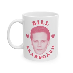 May include: White ceramic mug featuring a pink image of Bill Skarsgård, with the name "BILL" above and "SKARSGÅRD" below, in pink lettering. Two pink heart symbols flank the image. The mug has a white handle.