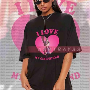 May include: Black unisex t-shirt with the text "I LOVE MY GIRLFRIEND" in pink. The shirt features a pink heart with a cartoon bunny character. The model is wearing sunglasses.