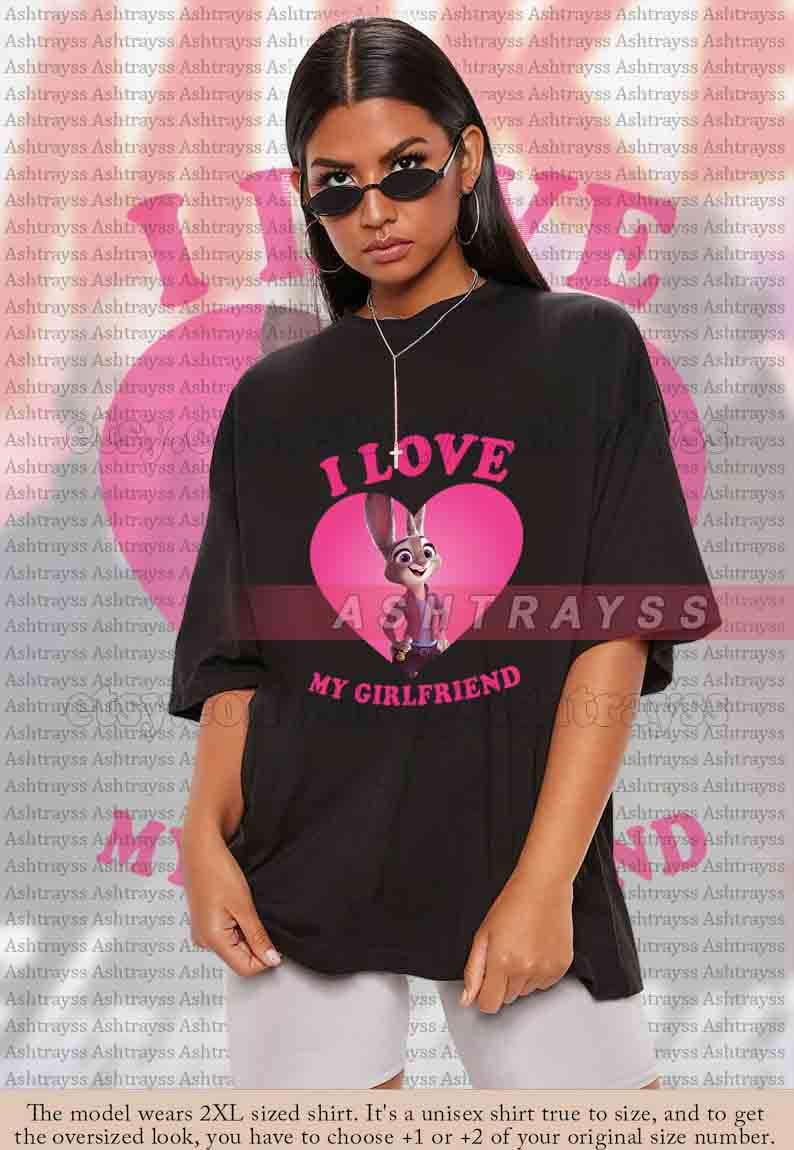 May include: Black unisex t-shirt with the text "I LOVE MY GIRLFRIEND" in pink. The shirt features a pink heart with a cartoon bunny character. The model is wearing sunglasses.