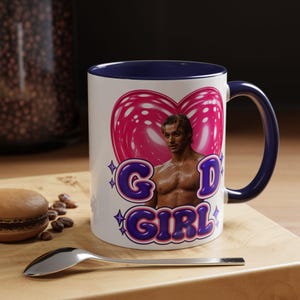 Peut inclure: Un mug en céramique blanche avec un intérieur et une anse bleu foncé. L'avant présente un graphique d'une personne torse nu à l'intérieur d'un cœur rose, avec le texte violet "GOD GIRL" et de petites étoiles entourant l'image.