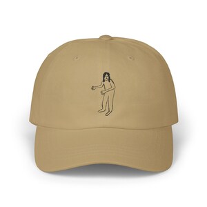 Peut inclure: Casquette de baseball beige avec un dessin au trait noir d'une personne aux bras tendus. La casquette a une visière incurvée et une conception à six panneaux. L'illustration est centrée sur le devant.