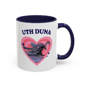 May include: A white ceramic mug with a dark blue handle and rim. The front features the text UTH DUNA in dark blue letters above a pink heart graphic containing a dark, winged creature illustration with small four-pointed star accents.
