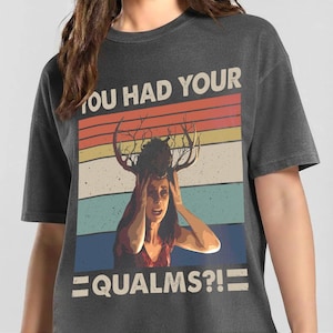 May include: A charcoal gray t-shirt featuring a graphic print of a person with deer antlers on their head. The design includes a retro-style sunset background with horizontal stripes and the text "YOU HAD YOUR QUALMS?!" in a bold, light-colored font.