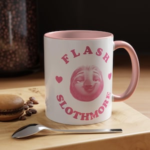 Puede incluir: Taza de cerámica blanca con interior y asa rosas. La taza presenta el texto "FLASH SLOTHMORE" en rosa, junto con una cara de perezoso rosa y corazones rosas. Una cuchara y un macaron están sobre una superficie de madera.