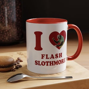 Puede incluir: Taza de cerámica blanca con interior y asa rojos. La taza presenta las palabras "I Flash Slothmore" en rojo, con un gráfico en forma de corazón que contiene un perezoso. Una cuchara y un pastelito están sobre una superficie de madera.