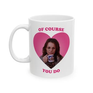 May include: White ceramic mug with a pink heart design and the text "OF COURSE YOU DO." The mug features an image of a woman holding a mug. The mug she is holding has a picture of a cabin.