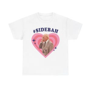 May include: A white t-shirt featuring a graphic print of a person in a suit covering their face with one hand, centered inside a pink heart with concentric rings. The text #SIDEBAH is printed in dark blue above the heart, with small dark blue stars.
