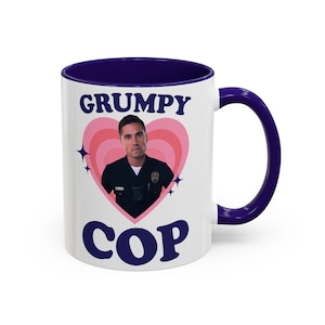 Peut inclure: Mug en céramique blanche avec intérieur et anse bleu marine. Le mug présente les mots "GRUMPY COP" en bleu marine, avec une photo d'un homme en uniforme de police à l'intérieur d'un cœur rose. Le design comprend des étoiles.