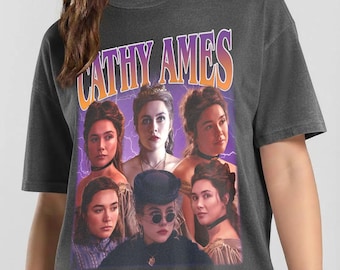May include: A charcoal gray short-sleeve t-shirt featuring a graphic print with the text CATHY AMES in orange and purple letters. The design includes five portraits of a woman with brown hair in various outfits against a purple background.