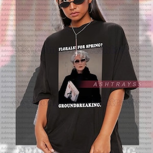 Könnte beinhalten: Schwarzes Unisex-T-Shirt mit einer Grafik einer Frau mit Sonnenbrille und dem Text "FLORALS FOR SPRING?" und "GROUNDBREAKING." Das Shirt ist übergroß und für verschiedene Körpertypen geeignet.