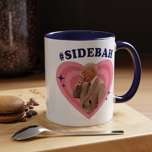 May include: A white ceramic mug with a dark blue interior and handle. The front features a pink heart graphic with concentric rings, a small star icon, and the text #SIDEBAH above a person in a tan suit covering their face with one hand.