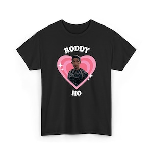 Könnte beinhalten: Ein schwarzes T-Shirt mit kurzen Ärmeln und einem Grafikdruck einer Person in einem rosa Herz mit konzentrischen Ringen. Der Text "RODDY" ist in Weiß über dem Herz und "HO" in Weiß unter dem Herz aufgedruckt, akzentuiert durch kleine weiße Sterne.