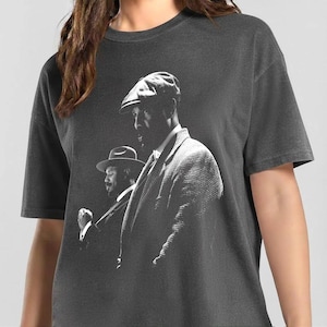 May include: A charcoal gray short-sleeve t-shirt featuring a black and white graphic print of two individuals wearing vintage-style hats and suits. The design shows one person in the foreground wearing a flat cap and another in the background.